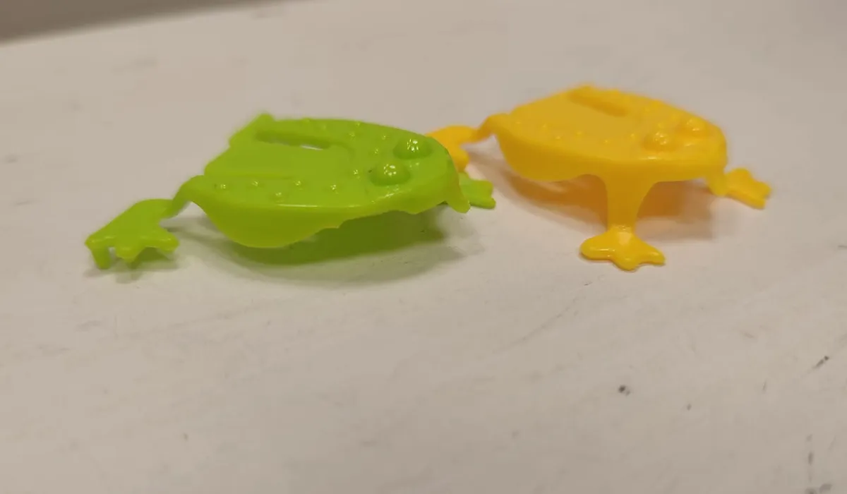Due piccole ranocchie salterine di plastica, una verde e una gialla, appoggiate su un tavolo bianco