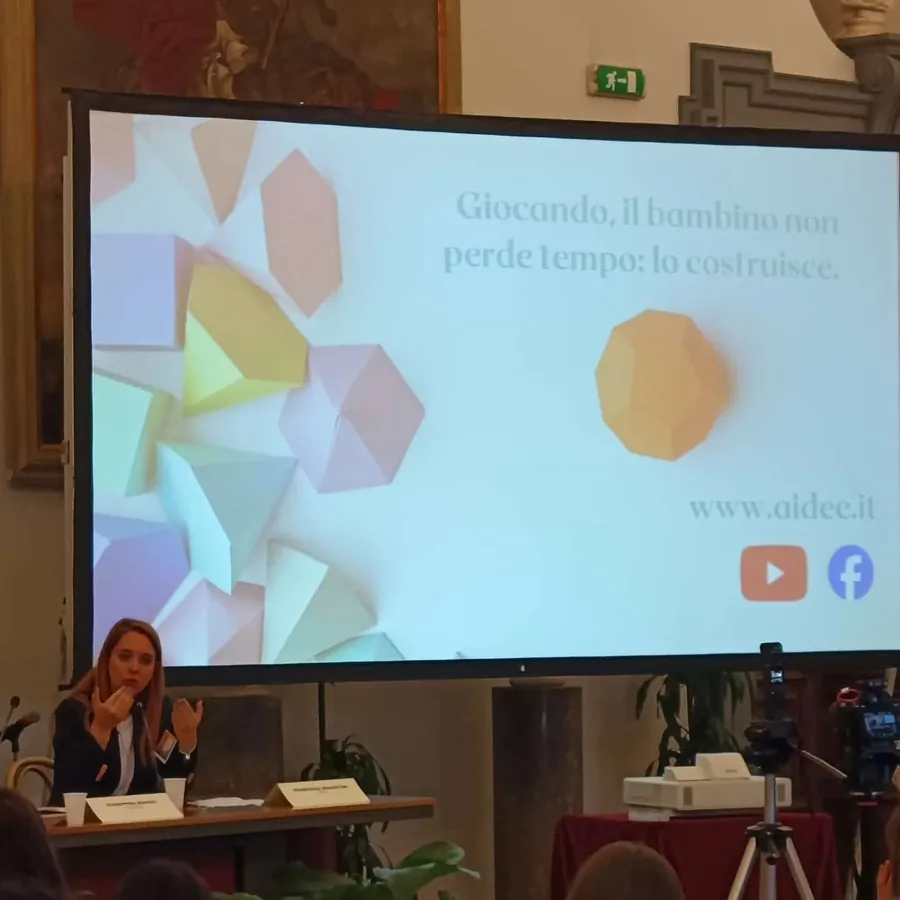 Francesca Magostini relatrice in una sala affrescata; sulla slide: «Giocando, il bambino non perde tempo: lo costruisce.»